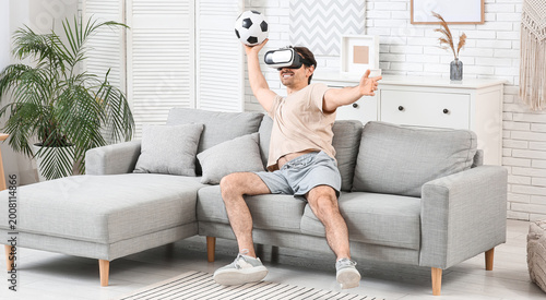 Sporty young man in VR glasses with soccer ball on sofa at home