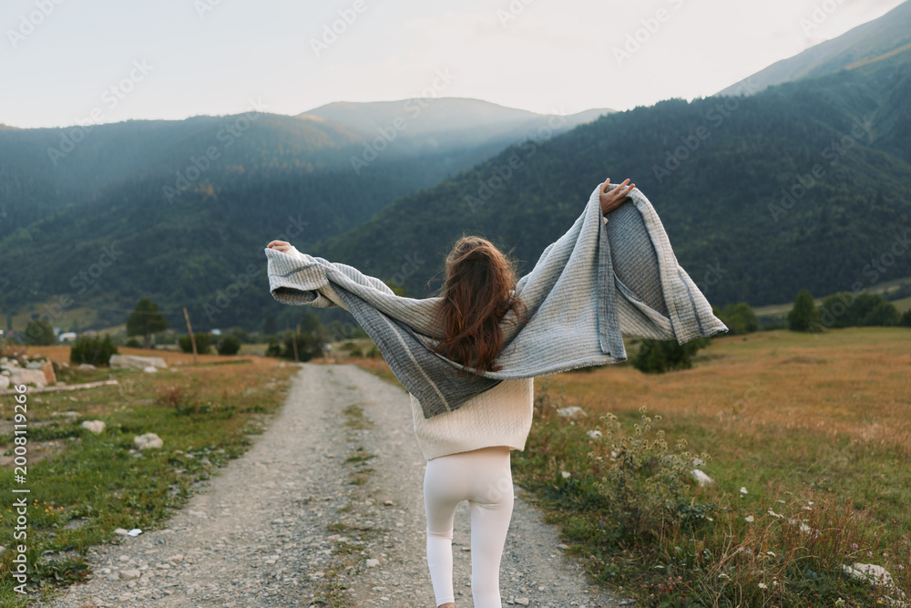 Fototapeta premium A young woman walks along a rural path through a sunlit valley, arms raised as a knitted shawl billows behind her, expressing freedom and outdoor lifestyle.