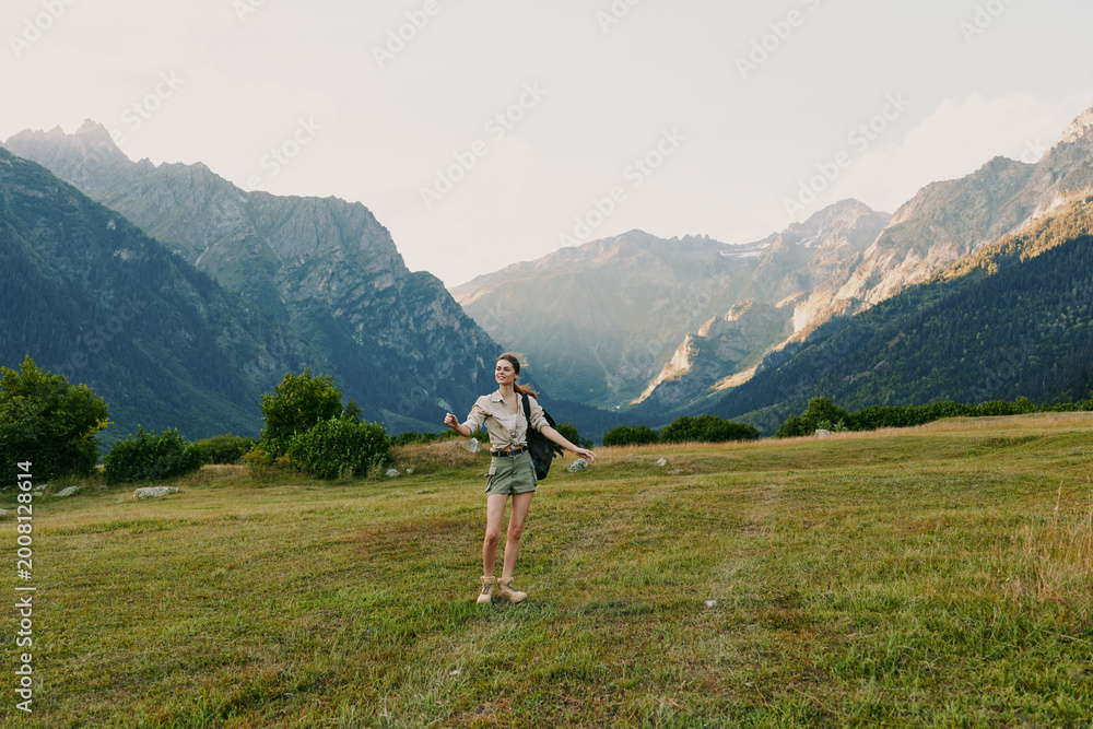 Fototapeta premium Young woman with long hair walks across a green meadow surrounded by mountains during a sunny day, enjoying nature and outdoor adventure with a casual outfit.