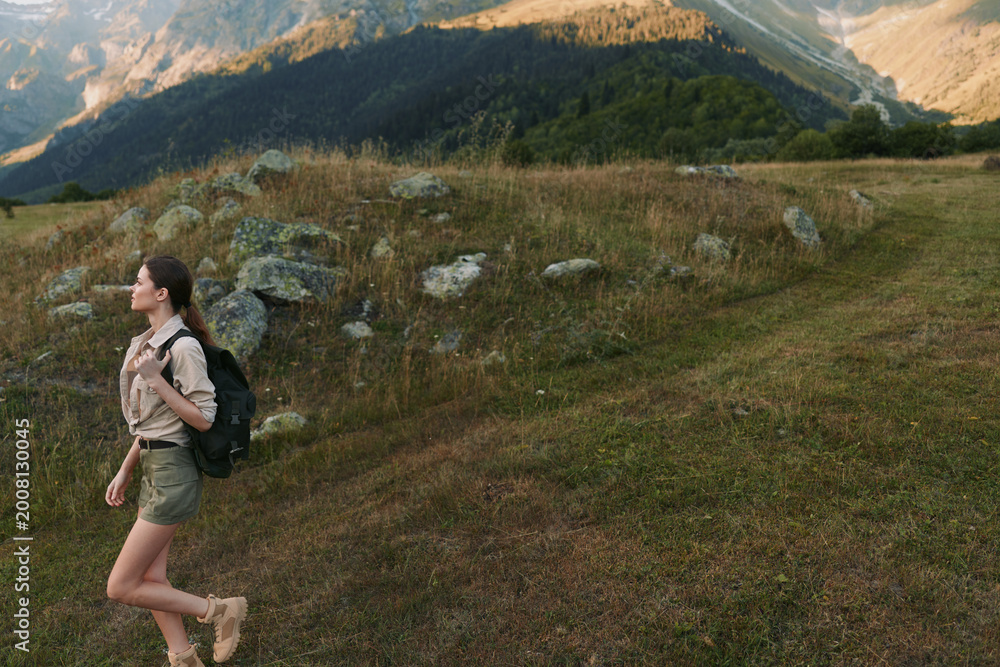 Naklejka premium Cheerful woman hiking outdoors with backpack, casual attire, grassy trail, mountain ridge in background, natural light, energetic lifestyle, scenic nature moment outdoors