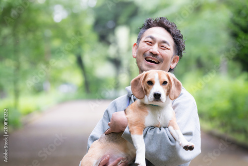 新緑背景で愛犬のビーグルを抱く男性の笑顔、公園で過ごす幸せなお散歩時間のカメラ目線クローズアップ