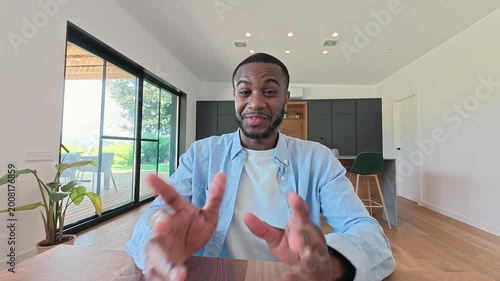 A young African American man smiles and gestures enthusiastically during a video call from his bright, modern home. He appears engaged and happy, possibly sharing ideas or having a friendly conversa