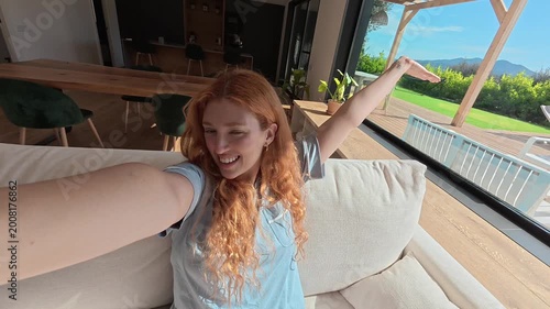 A cheerful young woman with vibrant red hair sits comfortably on a sofa, smiling and gesturing while recording a selfie video in her spacious, sunlit home.