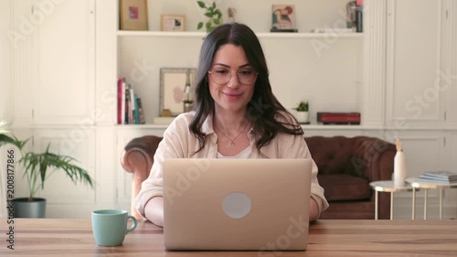 A cheerful young woman in glasses smiles confidently while working on her laptop at a home office desk. This scene embodies modern remote work and digital communication, reflecting productivity and 