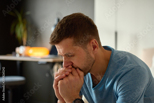 Photography Worried man sits indoors with clasped hands near his mouth and thoughtful expression in a home interior