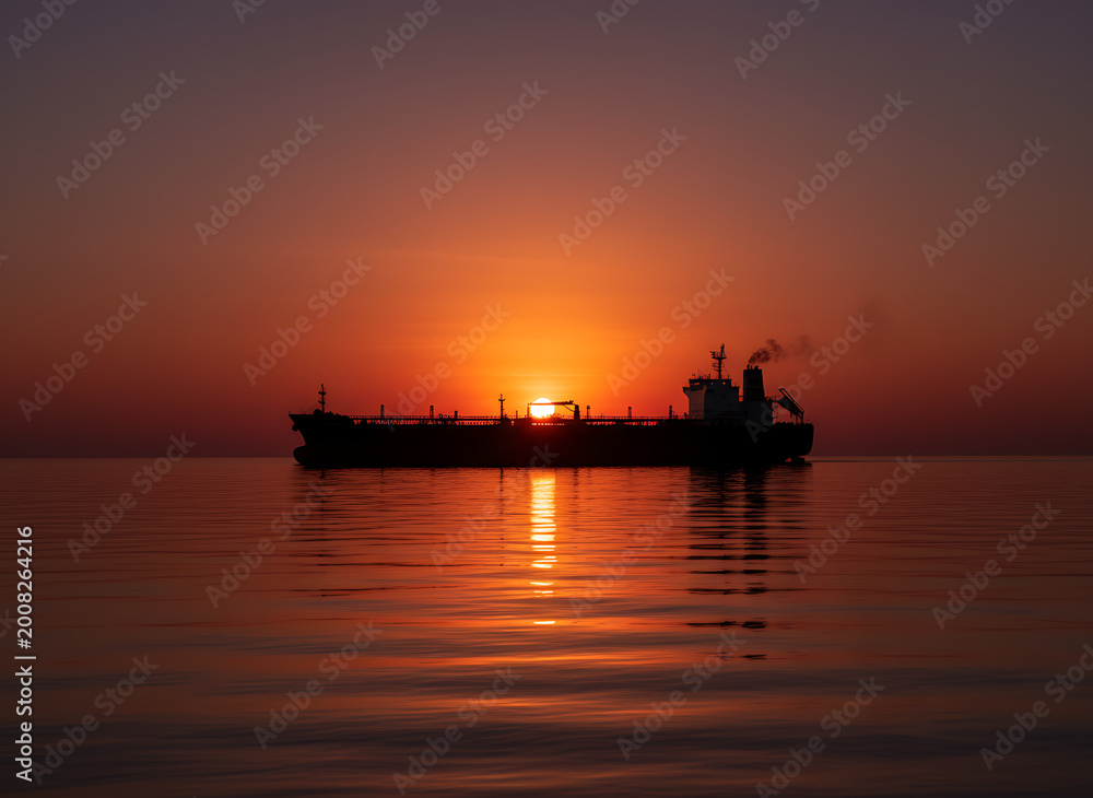 Fototapeta premium Crude oil tanker sailing across calm waters at vibrant orange sunset with reflection