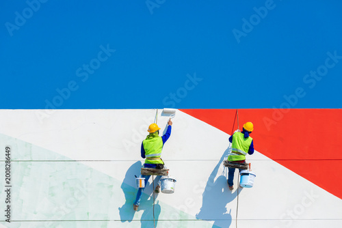 Workers paint a high building using a ladder, reflecting the risks of working at height, where safety, balance, and protective equipment are essential to prevent accidents.