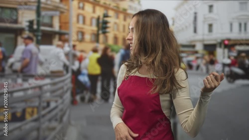 Hispanic woman in red apron gestures with hand, laughing and smiling while standing near a crosswalk on a busy city street; carefree joy.