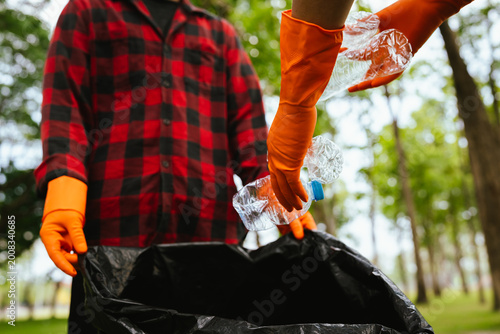 Photos Person wearing gloves placing a plastic bottle into a trash bag in a park, highlighting recycling, environmental cleanup, and responsible waste management outdoors