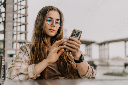 Smartphone woman outdoors using mobile device while sitting in outdoor cafe wearing stylish glasses and plaid shirt