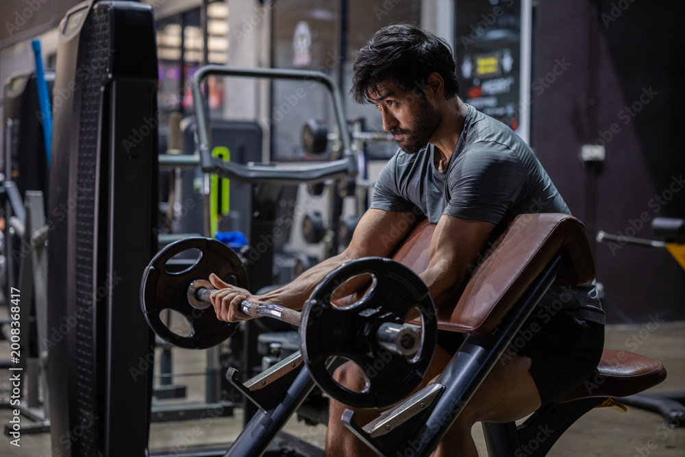 Naklejka premium Barbell bicep curl gym fitness strength training Young athletic man doing barbell bicep curl preacher bench modern gym, focused expression, muscular arm workout with weight plate equipment