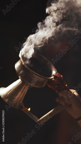 Varanasi, Uttar Pradesh, India. Man makes movements with a cup with smoking incense on the Ganga Maha Aarti ceremony. Brahman priest wearing red and yellow dress doing traditional ganga aarti at ghat
