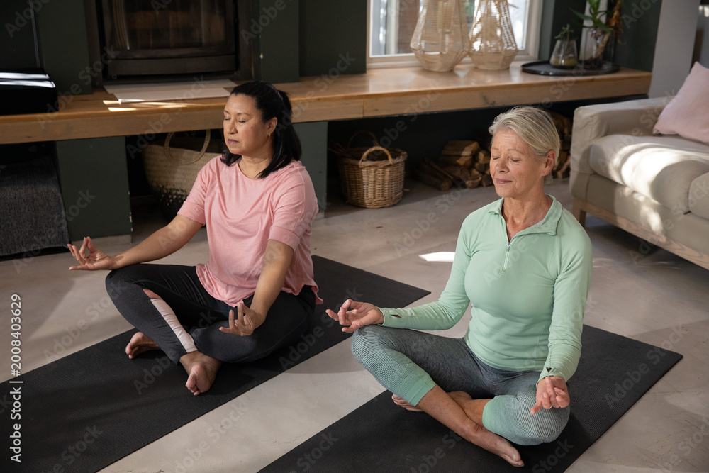 Fototapeta premium Female friends meditating cross-legged on black yoga mats in living room near wooden bench