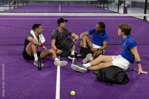 Diverse male teammates sitting on purple padel court near net, in blue shirts and holding paddles
