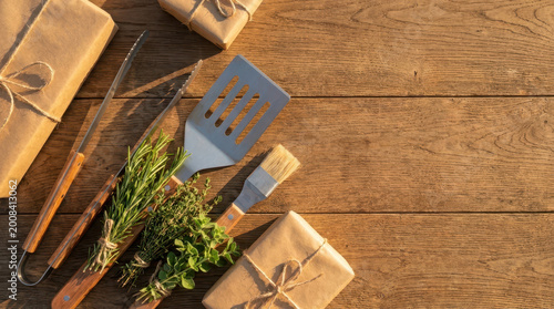Grilling tool Spatula Tongs Basting brush Herb bundle Rosemary Thyme Oregano Wrapped gift Brown paper Twine Wooden table Holidays Celebrations Cooking Barbecue Kitchen utensil Rustic Natural