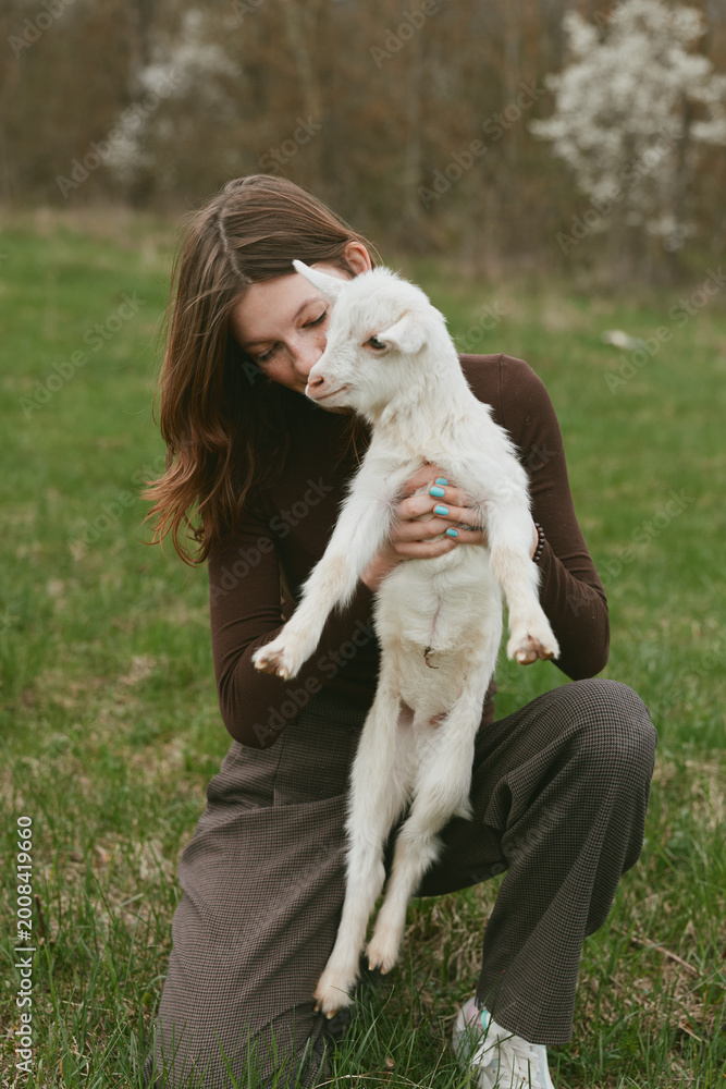 Fototapeta premium A woman gently holds a young goat in a grassy meadow, capturing a tender lifestyle moment of affection and care between human and animal in a natural rural setting.