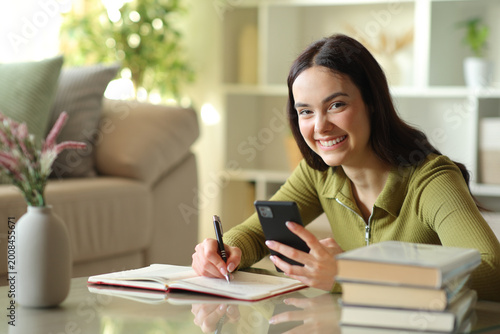 Happy young woman writing in agenda holding phone looks at you