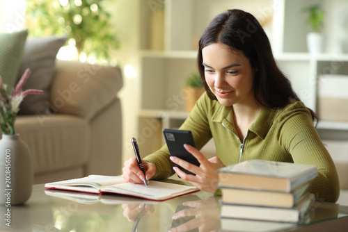 Canvas Print Satisfied young woman checking cell phone and writing in agenda