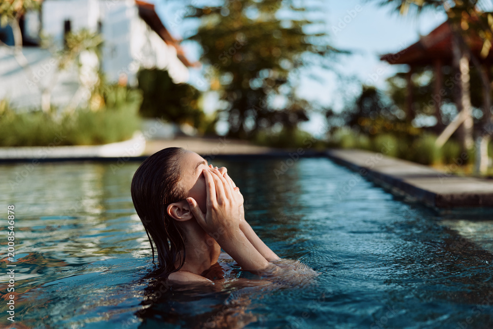 Fototapeta premium Woman in a pool, hands over her face, enjoying relaxation and self care by clear water and lush greenery, warm lighting, tranquil lifestyle moment in a sunny outdoor setting with calm reflections.