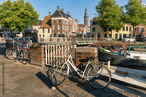 Bicycles parked beside canal in charming European town, with historic buildings, bridge, and trees under warm sunlight creating scenic urban atmosphere