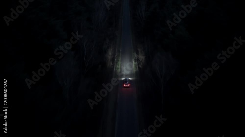 Car driving along straight road in dense forest at night filmed from drone with tracking movement and visible headlights
