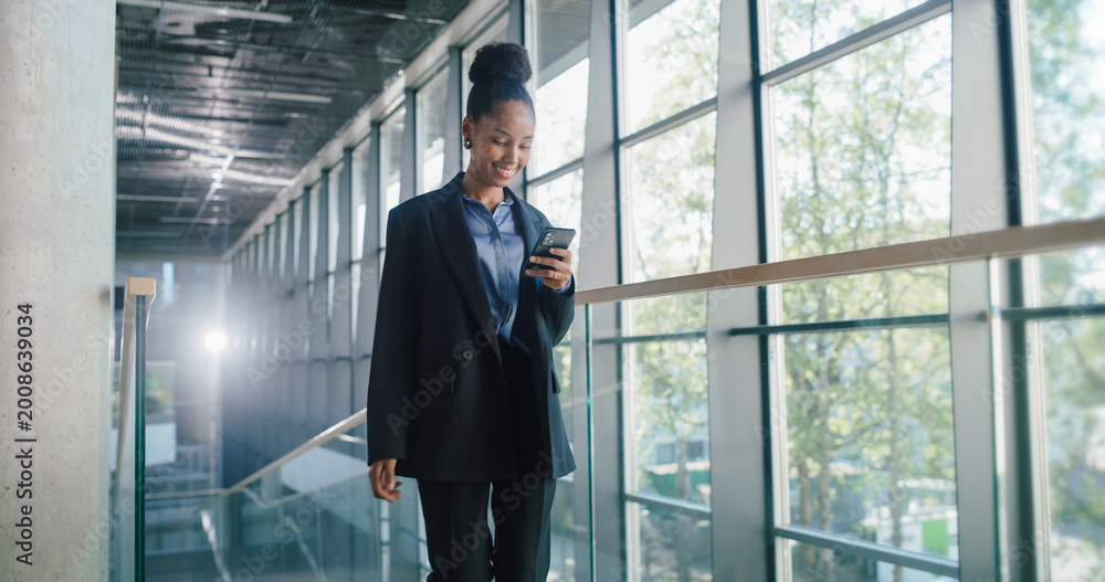 Fototapeta premium Young Businesswoman Walks Through a Bright Glass Office Hallway, Checking Her Smartphone to Track Sales Pipeline, Log Client Updates, Adjust Revenue Forecasts, and Coordinate Cross-functional Tasks