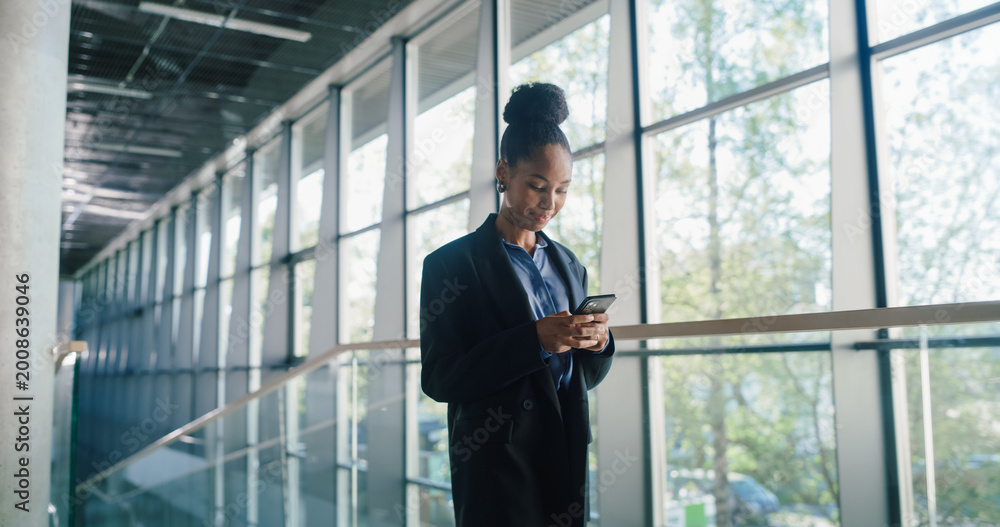 Fototapeta premium Young Businesswoman Checks Her Smartphone in a Bright Glass Office Corridor, Reviewing Project Timelines, Confirming Meetings, and Sending Quick Status Updates to Keep Team Communication
