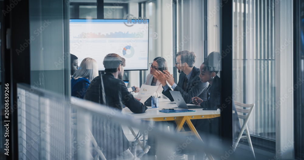 Obraz premium Business Leaders Sit Around a Conference Table Reviewing Charts on a Large Screen, Debating Results, Setting Priorities, and Planning Next Steps While Using Laptops, Printed Reports, and Notes During
