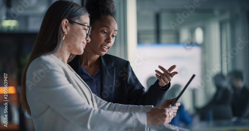 Fotografía Female Business Professionals Stand in a Modern Office Hallway Reviewing a Digital Tablet, Updating Sales Pipelines, Refining Customer Journeys, Planning Product Launches, and Aligning Revenue Target