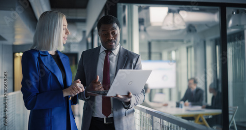 Two Coworkers Stand in a Glass Office Hallway Reviewing Sales Dashboards on a Laptop, Aligning Revenue Targets, Planning Client Follow-up Tasks, and Coordinating Next Steps While a Strategy Workshop 