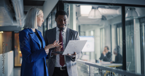 Coworkers Review Project Metrics on a Laptop in a Modern Office Hallway, Align Priorities, Clarify Tasks, and Plan Next Actions While a Business Meeting Continues in a Glass Conference Room