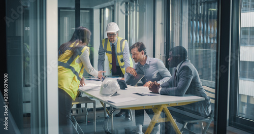 Executives and Construction Consultants Meet in a Glass Office to Review Blueprints, Align Budgets, Set Milestones, and Plan Risk, Compliance, and Stakeholder Communication Strategies for Business
