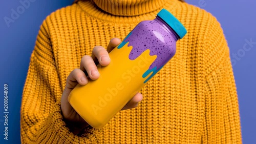 Female holding colorful drink bottle with purple and yellow design, wearing a cozy yellow sweater against a vibrant blue background in a studio setting