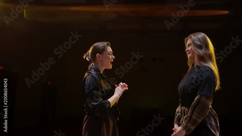 Two women rehearsing expressive dialogue under warm stage lights, practicing performance gestures and emotional delivery in a dramatic theater setting