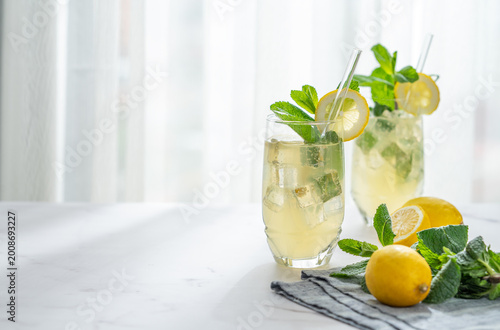 A summer drink with lemon, mint, and ice in a two glasses against a sunny window background.