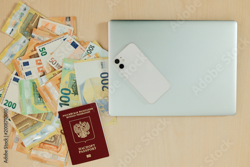 Russian passport lies on euro banknotes beside a laptop and smartphone on a desk. Concept of relocation from Russia, remote work setup and financial planning for moving abroad.