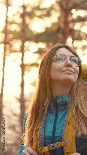A woman stands in a forest, gazing upward at the trees. The sunset casts a warm glow on her, enhancing her contemplative expression in the serene forest.