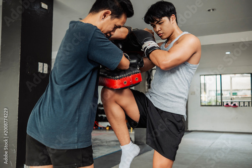 Bild auf Leinwand Athletic young man performs a powerful Muay Thai right knee strike on Thai pads held by coach during training