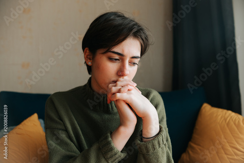 Canvastavla A young adult with short hair sits on a sofa with hands clasped together, appearing deep in thought