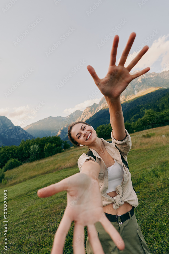 Fototapeta premium A woman in casual outdoor wear extends her hand toward the camera in a mountain meadow, smiling and enjoying a sunny day of active living and exploration in fresh air and wide open sky.