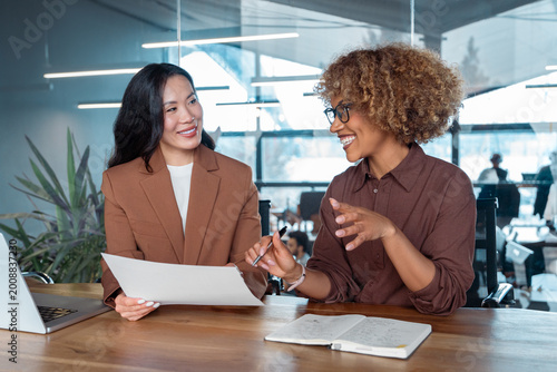 Foto Two businesswomen collaborating at modern office desk smiling during meeting with laptop and documents