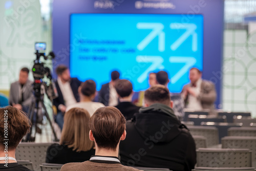 Audience watching panel presentation at business conference, camera recording, blurred stage, attendees seated, networking and learning atmosphere.