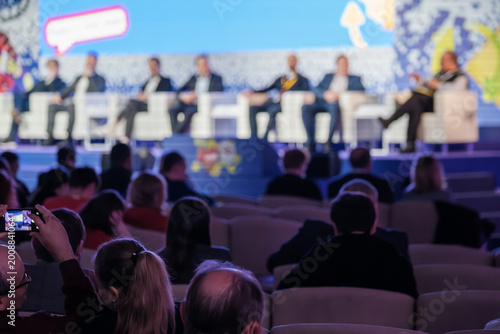 Fotografie Business conference panel on stage, audience watching and recording, blurred speakers and colorful backdrop, professional event atmosphere
