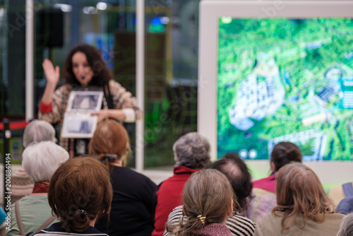 Group of older adults listening to presenter during guided tour, digital map screen in background, community education, social engagement, learning.