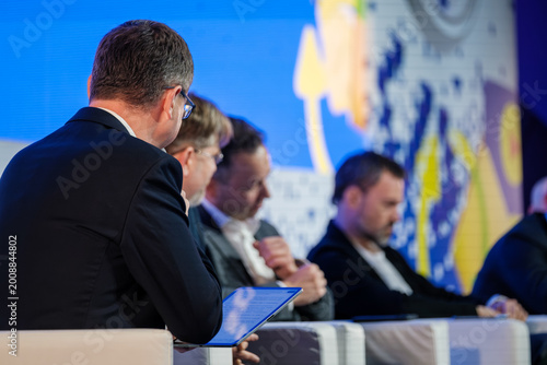 Business conference panel with executives using tablet, discussing strategy and networking on stage.