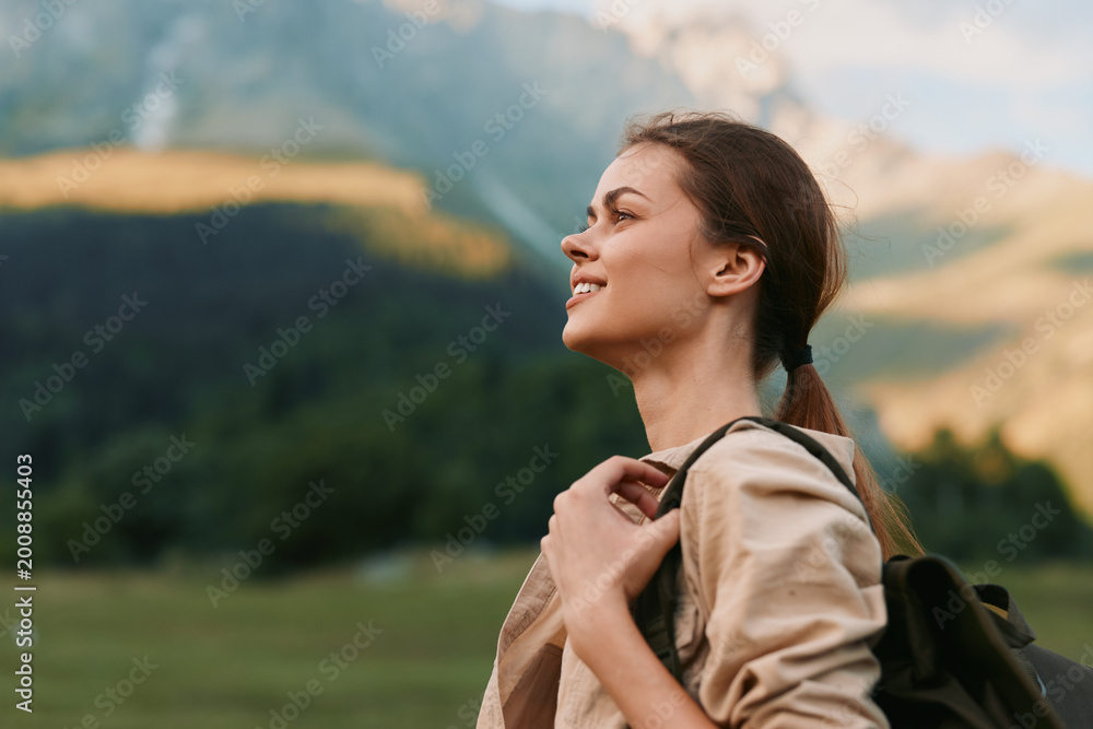 Fototapeta premium Woman traveler smiles while wearing a backpack in a meadow, with distant mountains, embodying outdoor lifestyle and exploration on a bright calm day. Scenic backdrop boosts mood.