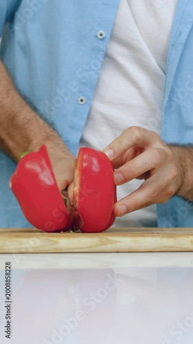 Preparation for family dinner concept where male hands holding sharp knife and cutting in half big fresh red pepper on wooden board at kitchen table