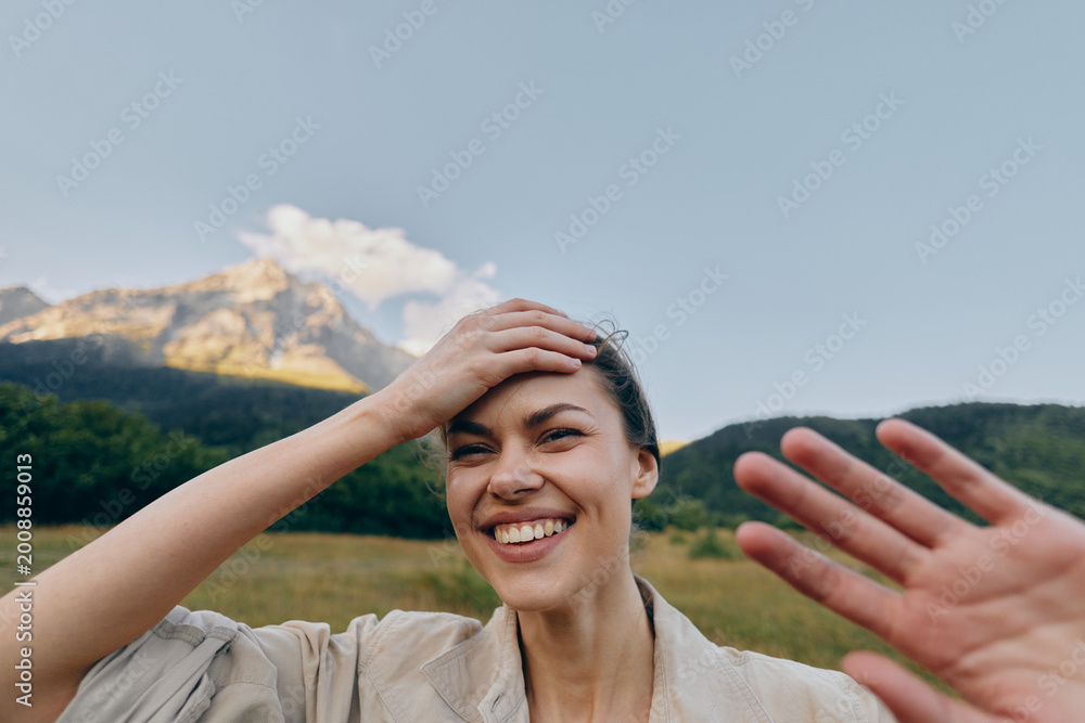 Obraz premium Cheerful woman outdoors, smiling with hand on head, sunlit moment, relaxed posture, natural colors, meadow and mountains in the background, gentle breeze, blue sky