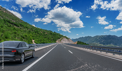 Εκτύπωση καμβά A black sedan drives along a modern asphalt highway winding through a lush green mountainous landscape under a bright blue sky with white clouds