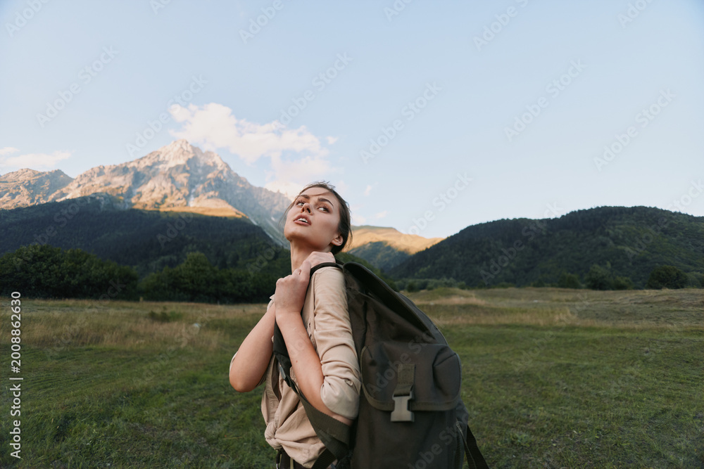 Fototapeta premium Young woman with a backpack gazes upward in an open meadow, mountains in the distance. Outdoor lifestyle, exploration, and self discovery through hiking journey.